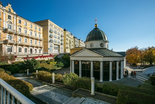 Pavillon with healing spring, Krizovy pramen, with white collumns and grey roof, yellow houses in background, Marienbad, Czech Republic, Europe, sunny day, clear blue sky, green bushes