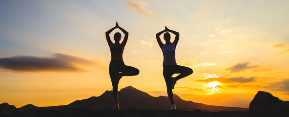Silhouette of young womans practicing yoga or pilates at sunset or sunrise in beautiful mountain location.