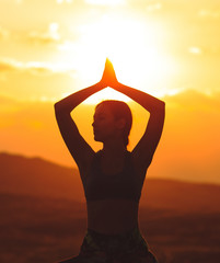 Silhouette of young woman practicing yoga or pilates at sunset or sunrise in beautiful mountain location.