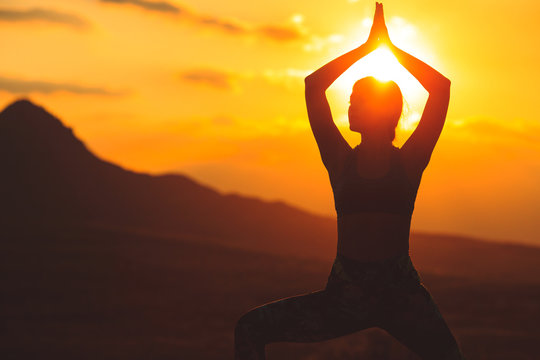 Silhouette of young woman practicing yoga or pilates at sunset or sunrise in beautiful mountain location.
