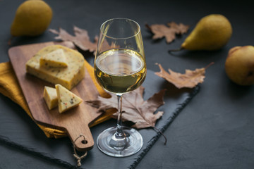 A glass of white wine served with cheese in a cutting board on dark background