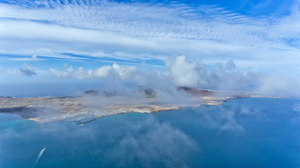Aerial view of La Graciosa island with fishing village, marina, volcano through clouds, north of Lanzarote, Canary Islands, Spain .