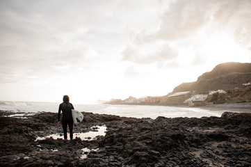 Rear view brunette boy standing in the black swimsuit with a white surf in his hands on the shore