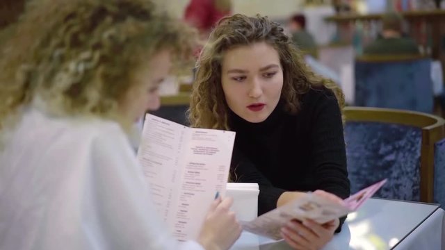 two pretty adult sisters are reading menu in restaurant and discussing, deciding what to order