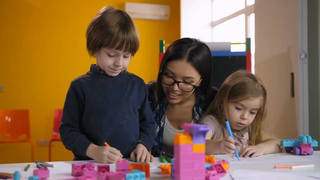 Positive attractive female asian teacher helping diverse preschool kids to draw at class in kindergarten. Smiling teacher and pupils working at the desk together during studying in preschool class.