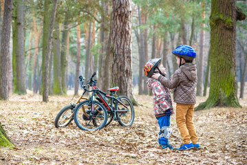 Plakat Two brothers preparing for bicycle riding in spring or autumn forest park. Older kid helping sibling to wear helmet. Safety and protection concept. Happy boys best friends having good time together.