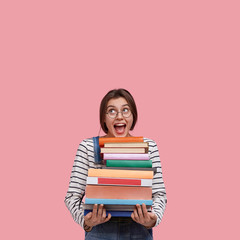 Studio shot of joyful European woman wears round spectacles, holds many books, opens mouth from excitement, poses against pink background with free space for your advertisement or promotion.
