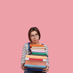Indoor shot of contemplative dissatisfied lady carries piles of books, wears optical glasses, looks upwards, isolated over pink background with free space for your advertisement or promotional content