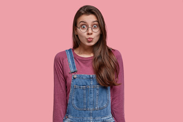 Pleasant looking brunette woman pouts lips, makes grimace at camera, wears purple jumper and denim overalls, wants to kiss someone, isolated over pink studio wall. People and facial expressions