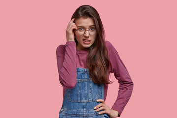 Indoor shot of pleasant looking young woman gazes in bewilderment, keeps one hand on head, other on waist, wears denim overalls, poses over pink studio wall. Negative facial expressions concept