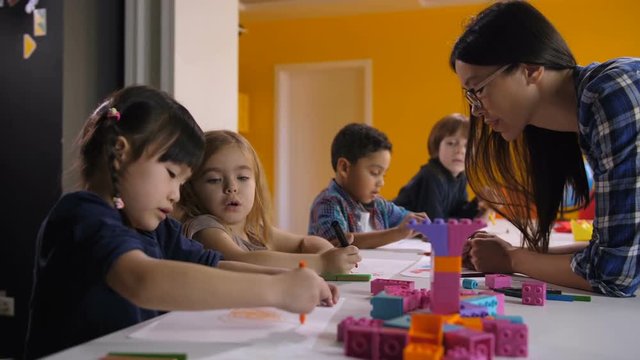 Cute Diverse Multi Ethnic Kids With Asian Teacher Drawing At Preschool Class. Positive Mixed Race Female Teacher Helping Multiracial Chilrdren With Drawing During Art Lesson In Kindergarten.