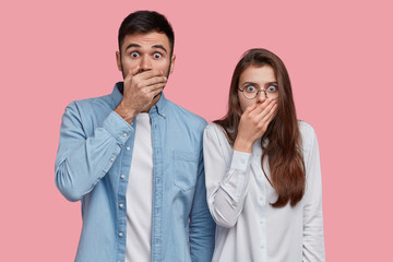 Photo of scared speechless lady and man cover mouthes with palms, have stupefied expressions, dressed in elegant shirt, stand next to each other over pink studio wall. People, reaction and feeling