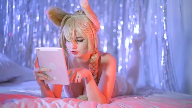 alone blonde woman is dressed in erotic lingerie and funny bunny ears is laying on bed with tablet