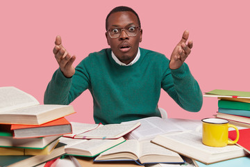 Photo of shocked black man raises hands in bewilderment, has wide opened eyes, dressed in green jumper, translates foreign article, uses many thick dictionaries, drinks hot tea, poses at desktop