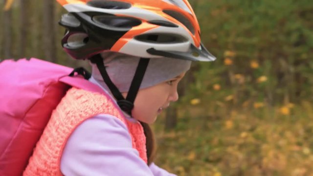 One caucasian children rides bike road in autumn park. Little girl riding black orange cycle in forest. Kid goes do bicycle sports. Biker motion ride with backpack and helmet. Mountain bike hardtail.