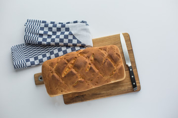 Freshly baked whole loaf of bread on a wooden chopping board, kitchen towel on the side, knife lying on a wooden board, top view.
