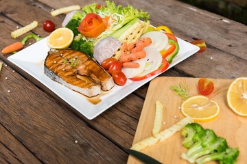 Salmon steak on a white plate with carrots, chili peppers, taro, tomatoes, grilled baby corn and other grilled vegetables.