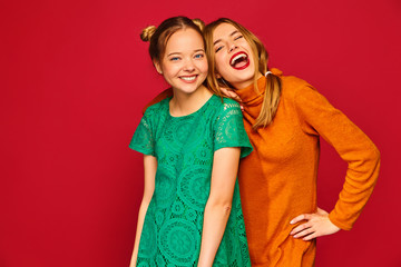Two young beautiful blond smiling hipster girls posing in trendy summer clothes. Carefree women isolated on red background. Positive models going crazy and hugging