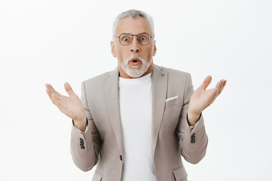 Waist-up Shot Of Shocked And Stunned Old Man Raising Hands In Surprise Folding Lips And Gasping Popping Eyes Confused And Questionned Cannot Undertand What Happening Posing In Suit And Glasses