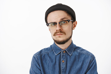 Doubtful and uncertain serious-looking bossy male entrepreneur in black beanie shirt and glasses raising one eyebrow in hesitation and disbelief standing unemotive and suspicious over gray background