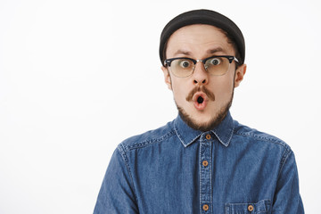Waist-up shot of charmed surprised handsome creative male artist in black beanie and transparent glasses folding lips and holding breath from amazement and thrill reacting to awesome news