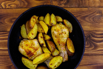 Baked chicken drumsticks with potatoes in in cast-iron frying pan on wooden table. Top view