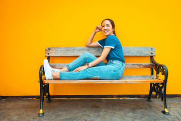 portrait of fashion hipster girl sit on wood bench beside yellow well  with posing relaxing smile by wearing blue shirt and blue jeans