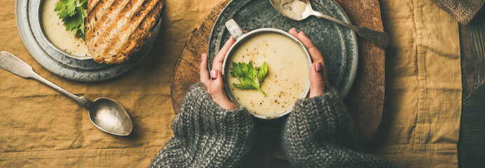 Autumn, Winter home dinner. Flat-lay of Fall warming celery cream soup, grilled bread and female hands over linen tablecloth, top view, wide composition. Comfort food, healthy and slow food concept - obrazy, fototapety, plakaty