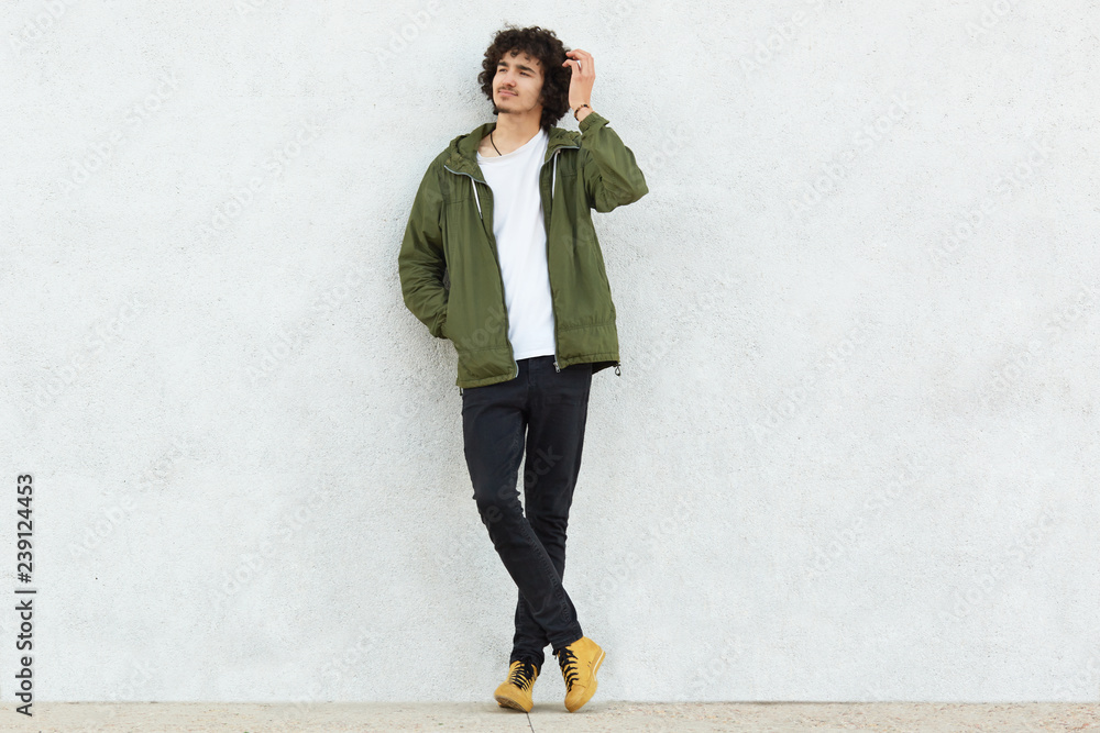 Wall mural Photo of attractive curly man with thoughtful expression, scratches head, dressed in fashionable clothes, poses against white studio wall, feels lonely thinks about something. People and style concept