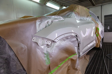 Painting the car body parts of the driver's door and the white wing in the spray booth in the body repair shop; other elements are covered with film and paper from splashing.