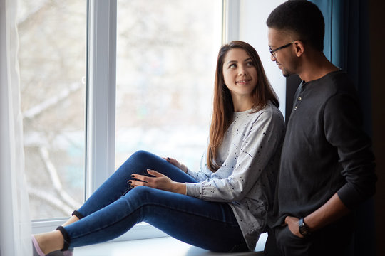 Photo of pleased interracial couple enjoys domestic atmosphere, pose near window in living room, have friendly talk, dressed in casual clothes, have good relationship. Mixed race woman and man