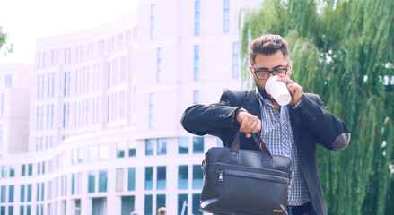 Portrait of a young man in a jacket with a beard and glasses walking down the street drinking coffee and looking at the clock.