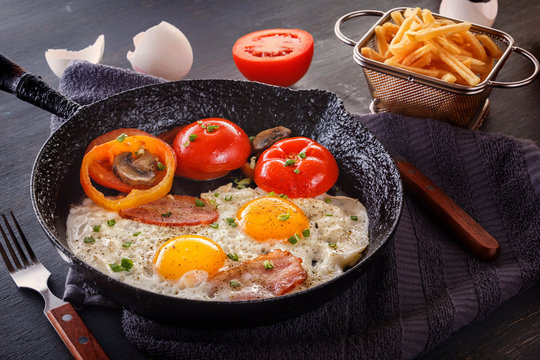 Fried eggs with bacon and tomatoes on an old cast-iron pan with french fries on a gray table. Close-up