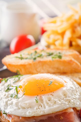 Fried egg with bacon in a black plate with fried pieces of bread, greens tomatoes, a jug of milk and French fries on a gray wooden table. Close-up
