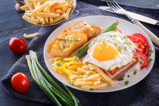 Fried egg with bacon in a white plate with fried pieces of bread, greens, tomatoes and french fries on a gray wooden table. Close-up