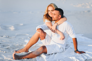 A bearded man and a blond woman hug sitting on the white sand and enjoy the sunset. Love in the desert newlyweds. The love story of fun and love people.