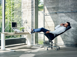 Junger Mann telefoniert entspannt in halbliegender Position auf Bürostuhl