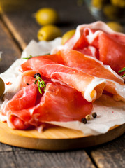 Closeup of thin slices of prosciutto with mixed olives and paprika on wooden cutting board