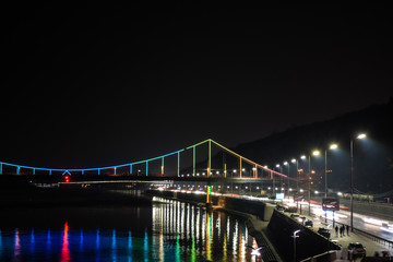 Night european city in colorful lights and reflection in water, Kyiv (Kiev) the capital of Ukraine. Pedestrian bridge across the Dnieper river and view from the river station, evening illumination