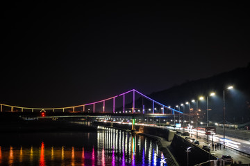 Fototapeta na wymiar Night european city in colorful lights and reflection in water, Kyiv (Kiev) the capital of Ukraine. Pedestrian bridge across the Dnieper river and view from the river station, evening illumination