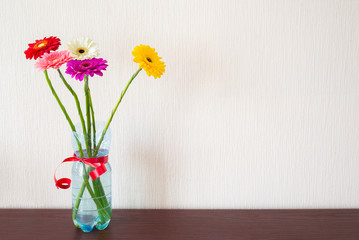 Five colorful Gerbera flowers in a plasctic water bottle with a red ribbon, on a wooden table