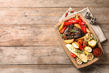 Board with barbecued steak, garnish and sauce on wooden background, top view. Space for text