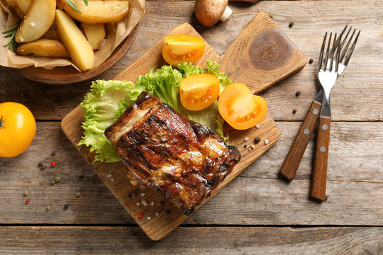 Board with barbecued ribs and garnish on wooden background, top view