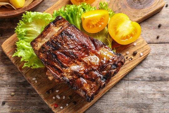 Board with barbecued ribs and garnish on wooden background, top view