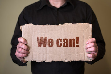 A man in a dark shirt holding a piece of cardboard with text We can