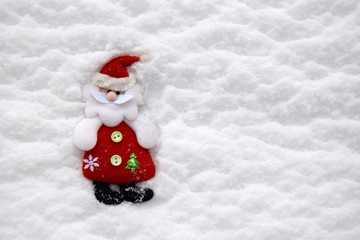 Christmas soft toy in the form of Santa Claus. The toy is located against the background of white natural snow. Santa Claus is dressed in a red fur coat, a cap and black valenoks