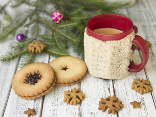 Winter morning, cup of coffee with knitted cup holders, gingerbread cookies with fir tree branches decorated with spruce balls on white wooden background.Breakfast. Christmas mood