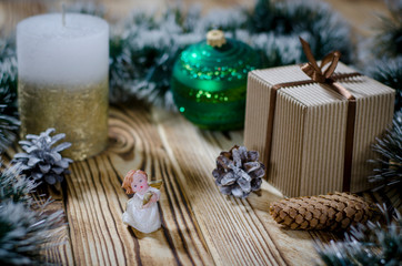 A gift lays on a wooden table next to a candle, cones and an angel against the background of Christmas decorations.