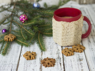 Winter morning, cup of coffee with knitted cup holders, gingerbread cookies with fir tree branches decorated with spruce balls on white wooden background.Breakfast. Christmas mood