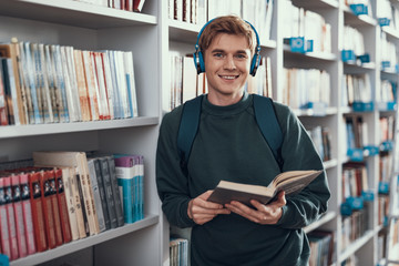 Handsome student standing near the bookcase and holding his book